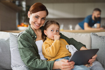 Mother with small daughter using tablet indoors at home, looking at camera, everyday life and home office with child concept.