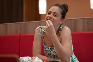 Muchacha joven adolescente de pelo oscuro con moño y vestido estampado comiendo en un restaurante con felicidad y risueño sentada en la mesa