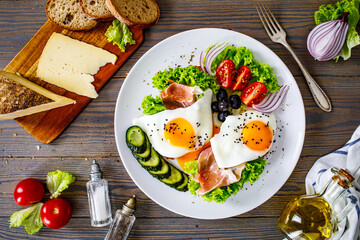 Sunny side up eggs on toasted bread and prosciutto di Parma  with fresh green vegetables, mini tomatoes and black olives on wooden table
