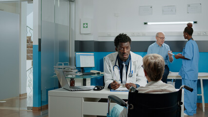 Elder patient with disability having checkup appointment with young doctor at medical clinic. Invalid old woman in wheelchair sitting at desk with medic while talking about healthcare problems