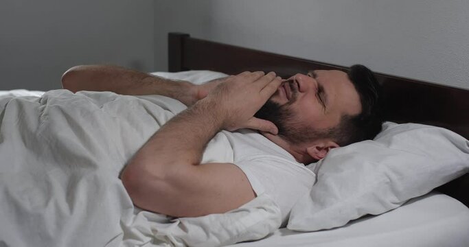 Close up  of caucasian bearded man lying in bed with white linens in light room suffering from toothache. Attractive guy suffers from dental  pain  holding face with hands   4K 