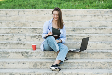 A happy and attractive Caucasian girl is sitting with a notebook on the steps and writing in a notebook. Distance working. Freelance concept