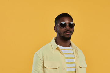 Fototapeta premium Minimal waist up portrait of trendy African-American man wearing sunglasses and looking at camera while standing against yellow background in studio, copy space