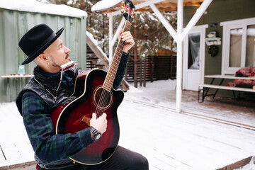 Cool looking musician in a hat and leather jacket playing guitar on snowy floor
