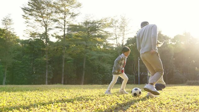 asian father playing soccer with son outdoors in park while mother and daughter watching from behind