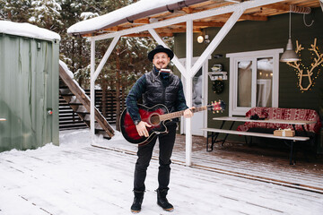 Musician in a hat and leather jacket standing in front of house on a snowy floor