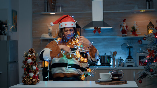 Smiling woman with santa hat tangled in christmas lights string while doing decor preparations in kitchen. Young person having fun indoors with festive decorations and ornaments