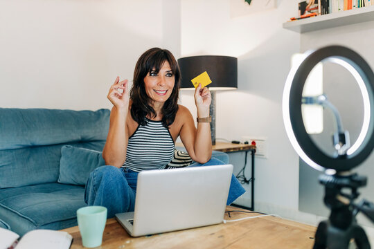 Smiling female vlogger with credit card filming through smart phone in living room
