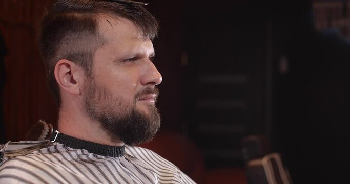 Close-up of a handsome Caucasian guy sitting in a hairdressing salon. An unrecognizable barber shaves a bearded man's head with a trimmer. 4K 50 fps slow motion 