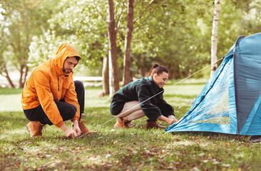 camping, tourism and travel concept - happy couple setting up tent outdoors