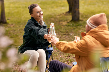 Naklejka premium camping, tourism and travel concept - happy couple drinking beer and eating sandwiches at tent camp