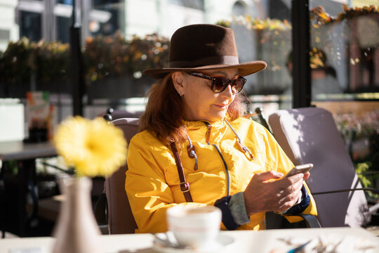 Beautiful happy mature senior woman in hat sunglasses and yellow jacket sitting in street cafe using smartphone
