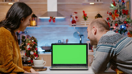 Festive couple looking at horizontal green screen on laptop for christmas eve celebration. People...