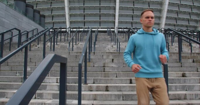 Front view tracking shot of sporty young man dressed in green hoodie and  brown pants jogging downstairs  against moden beautiful stadium. Caucasian guy running the stairway.
50 fps slow motion