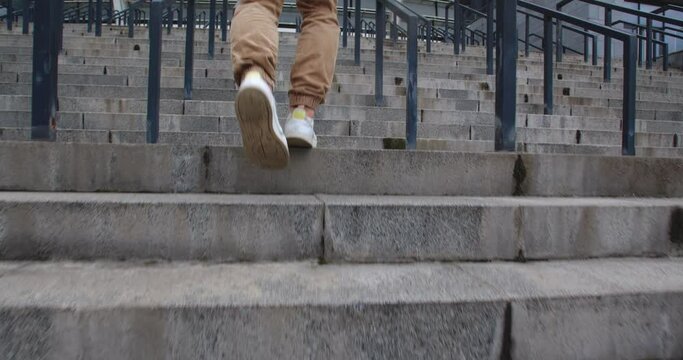 Rearview close up tracking shot of sporty young man dressed in green hoodie and  brown pants jogging upstairs  against moden beautiful stadium. Unrecognizable guy running the stairway.
