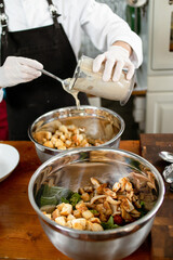 The chef is dressing the caesar salad with sauce. Ingredients in a bowl. Chopped vegetables and baked croutons. Food preparation technique. On a wooden table in rustic style