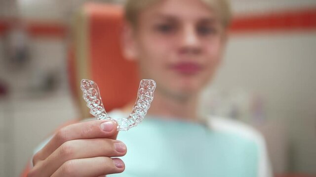 Teenager showing mouthguards and sitting while visiting orthodontist in clinic office spbd. Closeup view of caucasian boy holds transparent sample in hand and showes, looks with smile and sits in