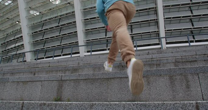 Rearview  of sporty young man dressed in green hoodie and  brown pants jogging upstairs  against moden beautiful stadium.  Unrecognizable guy running the stairway.
50 fps slow motion