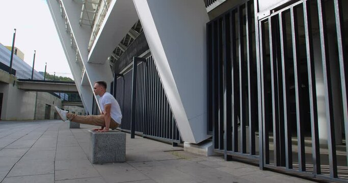 Professional acrobat performing trick on the street against backdrop of modern stadium. Caucasian guy  making a handstand with straddle split on a playground  50 fps slow motion
