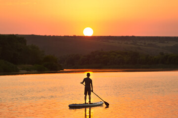 A man on a SUP on the river in the summer admires the sunset. Silhouette of a man on a sup on the...