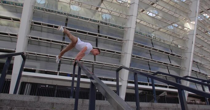 Professional acrobat performing dangerous trick on the railings against backdrop of modern stadium. Caucasian guy  making a handstand with straddle split on a narrow metal pipe
50 fps slow motion
