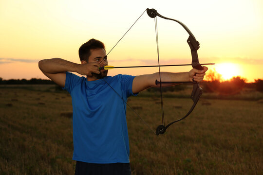 Man with bow and arrow practicing archery in field at sunset