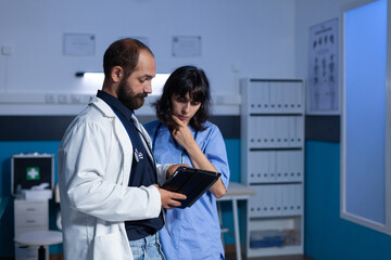 Fototapeta premium Doctor and nurse working late at night for healthcare using digital tablet. Medical assistant and medic doing teamwork at office for medicine. Woman and man looking at modern gadget