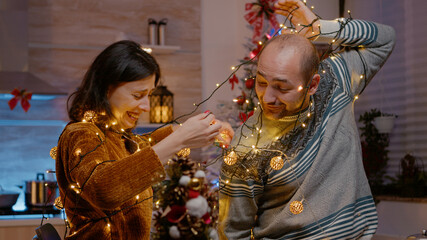 Couple laughing getting tangled in string of festive lights while decorating kitchen for christmas...