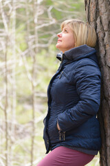 Woman at the trunk of a tree. Looks up. Blonde in a blue jacket. Cool weather. Tree bark.