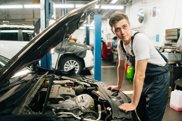 Side view of confident handsome professional male car mechanic in blue uniform standing in front of open hood, inspecting engine of car coming in for repair or maintenance, looking at camera.