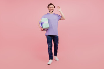 Full body photo of young guy happy positive smile hold gift box go wave hello isolated over pink color background