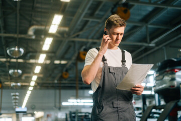 Low-angle view of focused handsome young mechanic male wearing uniform reading clipboard and...