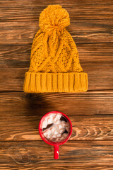 top view of yellow knitted hat near cup of cocoa with marshmallows on wooden surface