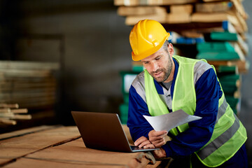 Mid adult foreman analyzing paperwork while working on laptop at distribution warehouse.