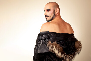 Portrait of bald made-up man with bare back wearing feather jacket. Image in low angle. Side view of caucasian drag man with beard.