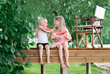 Two happy kids- little boy and girl eating watermelon on wooden terrace in home backyard