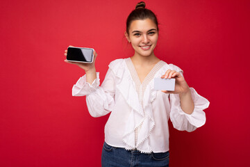 Naklejka premium Young beautiful european stylish brunette woman wearing white blouse and fashionable hat isolated over red background with positive sincere emotions. Simple and natural looking at the camera. Holding