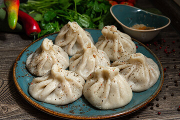 Georgian-style khinkali with meat on a handmade ceramic plate. Sprinkled with ground pepper. Leaves of parsley and coriander in the background. On a dark grey wooden kitchen table.