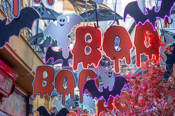 Halloween decoration. Hanging garlands. Bloody BOO text, white flying ghosts, black umbrella and bats outside of a store. Festive decorative lights calling for celebration. Costume party preparation