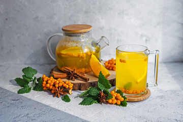 Still life of autumn hot tea from fresh sea buckthorn, mint and orange on a gray stone background. A healthy vegan drink. Medical. Sea buckthorn and mint branch. Health drink ingredients. Autumn. 