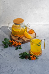 Still life of autumn hot tea from fresh sea buckthorn, mint and orange on a gray stone background. A healthy vegan drink. Medical. Sea buckthorn and mint branch. Health drink ingredients. Autumn. 