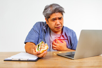 This middle-aged man eats a hamburger while sitting at work, laptop, computer, has chest pain,...