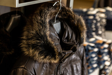 A black leather men's jacket with a puffy fur hood, collar hanging in a men's clothing store against blue jeans.