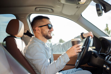 Portrait of a smiling businessman driving a car while drinking coffee. He is looking happy.