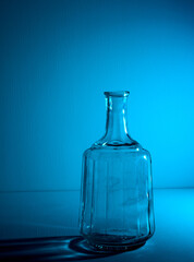 Glass empty open bottle with copy space. Graphic still life with light and shadow in blue light. The concept of glass containers, recyclable materials or alcoholism. Large creative vase. Open flask