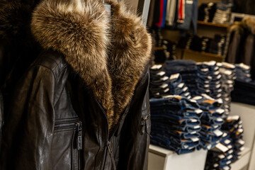 A black leather men's jacket with a puffy fur hood, collar hanging in a men's clothing store against blue jeans.