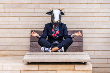 Businesswoman wearing bull mask practicing lotus position sitting on bench by wall