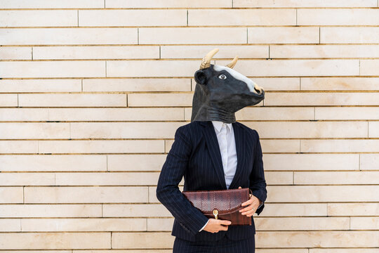 Female entrepreneur wearing bull mask holding purse while standing by wall