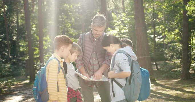 Caucasian man teacher standing in forest and examining route plan with her pupils. Male couch resting in wood with kids while having walk outdoor and looking at map. Tourism concept