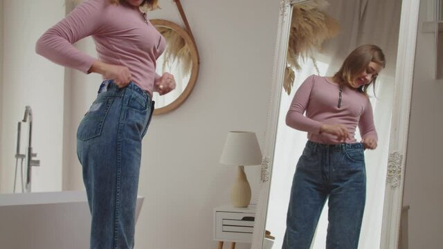 Midsection of charming happy slimming female looking at her reflection in big mirror with cheerful smile, checking successful weight loss program by wearing old oversized jeans in domestic room.